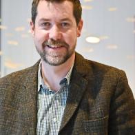 Mickey Green, a man in his 40s with short dark hair and a beard, wearing a tweed jacket and checked shirt