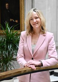 Katie Stewart, a white woman in her mid-40s, with shoulder-length blond hair, wearing a pale pink jacket and smiling at the camera