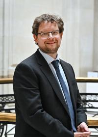 David Shepherd, a white man in his 40s, with mid-brown hair, beard and glasses, wearing a dark suit, pale blue shirt and dark blue tie