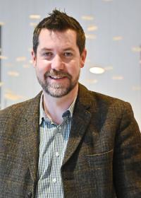 Mickey Green, a man in his 40s with short dark hair and a beard, wearing a tweed jacket and checked shirt